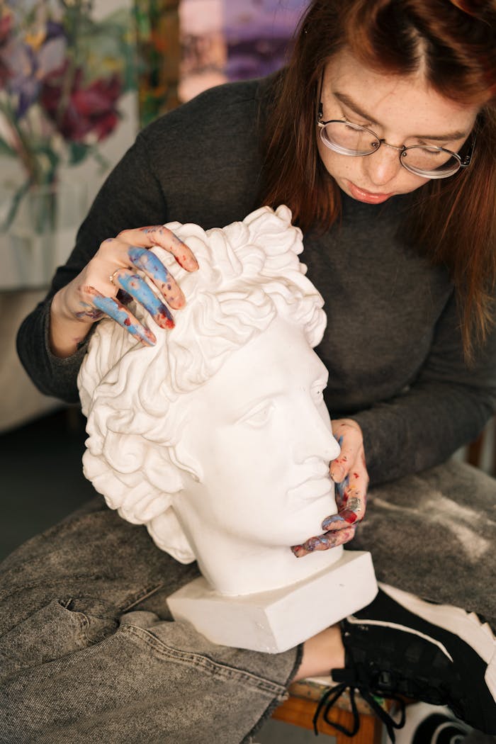A female artist in a studio meticulously works on a marble bust with paint-stained hands.